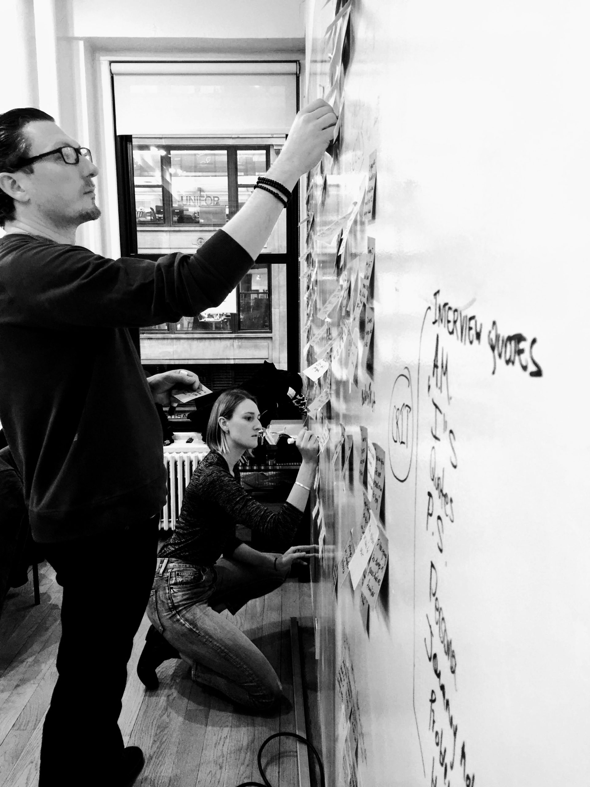 Black and white photo of 2 people affinity mapping, arranging post-it notes on white board.
